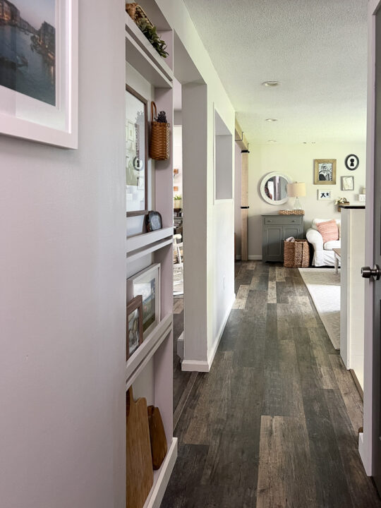 looking down the hall to the living room with shelf next to fridge. 